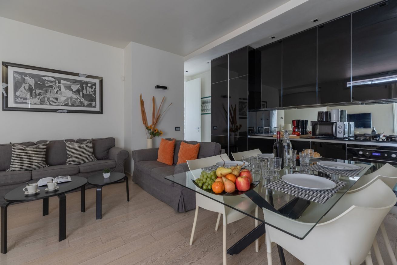 A modern living room with a grey sofa, a glass dining table set with fruits, and a kitchen with dark cabinets and appliances.