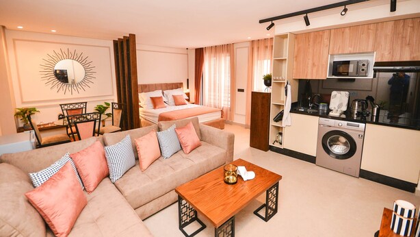 A modern hotel room with a sofa, a wooden coffee table, a bed, a kitchen area with a microwave and washing machine, and a large mirror on the wall.