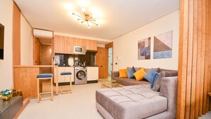 A modern living room with a grey sofa, wooden accents, a kitchen area with a microwave, and a washing machine.