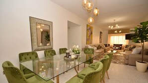 Een moderne woonkamer met een glazen eettafel, omringd door groene bekleden stoelen, een zithoek en een grote potplant.