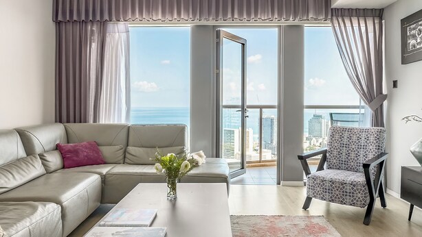 A modern living room with a sofa, a patterned armchair, and a coffee table. A large window offers a view of the cityscape. - High-Rise Sea-View Stay by Sea N' Rent (Tel Aviv)