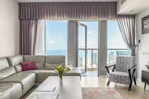 A modern living room with a sofa, a patterned armchair, and a coffee table. A large window offers a view of the cityscape. - High-Rise Sea-View Stay by Sea N' Rent (Tel Aviv)