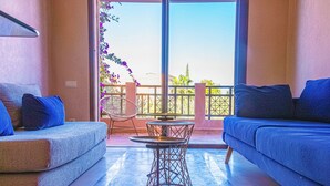 A modern living room with a sofa, a small table, and a chair. There is a balcony with a view of the sky and some plants. - Waky Club | Designer Flat (Aghouatim)