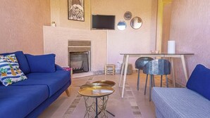A living room with a blue sofa, a small round table, a TV mounted on the wall, and a fireplace. - Waky Club | Designer Flat (Aghouatim)
