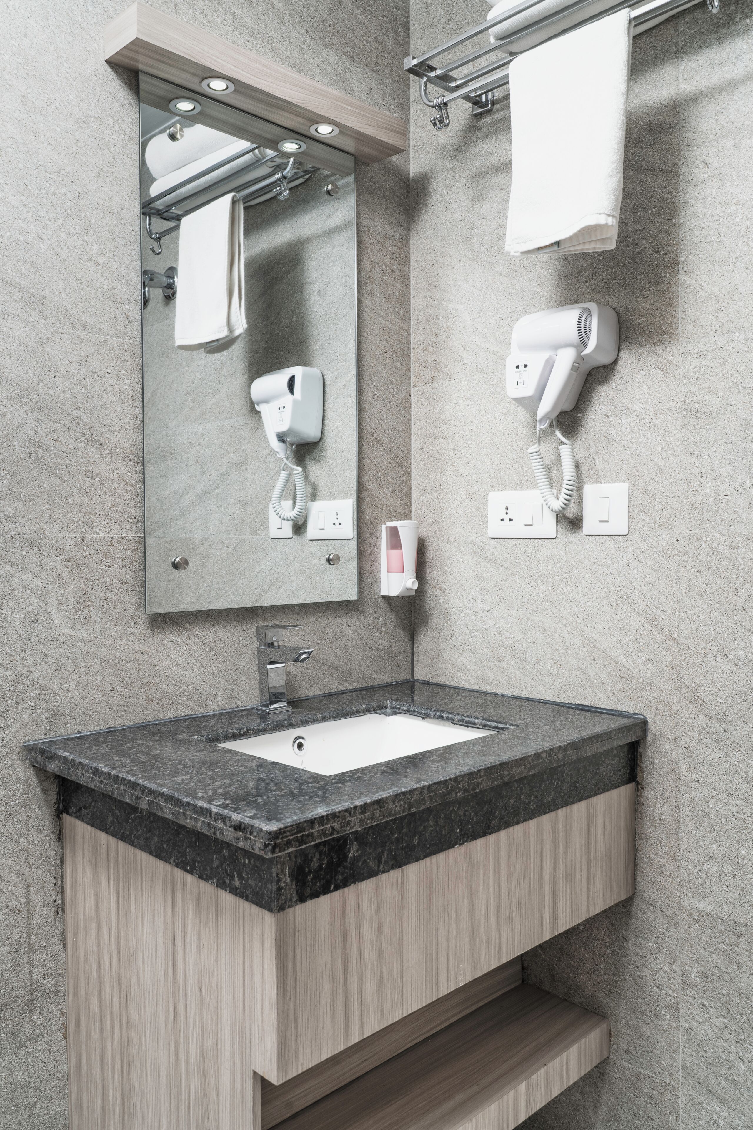 A modern bathroom with a granite countertop, a mirror, a towel rack, a hairdryer, and a soap dispenser.
