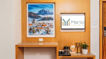 Un cuadro enmarco que muestra un pueblo montañoso, un letrero de 'Marlo Hotels' y una recepción con una cafetera y una planta en maceta.
