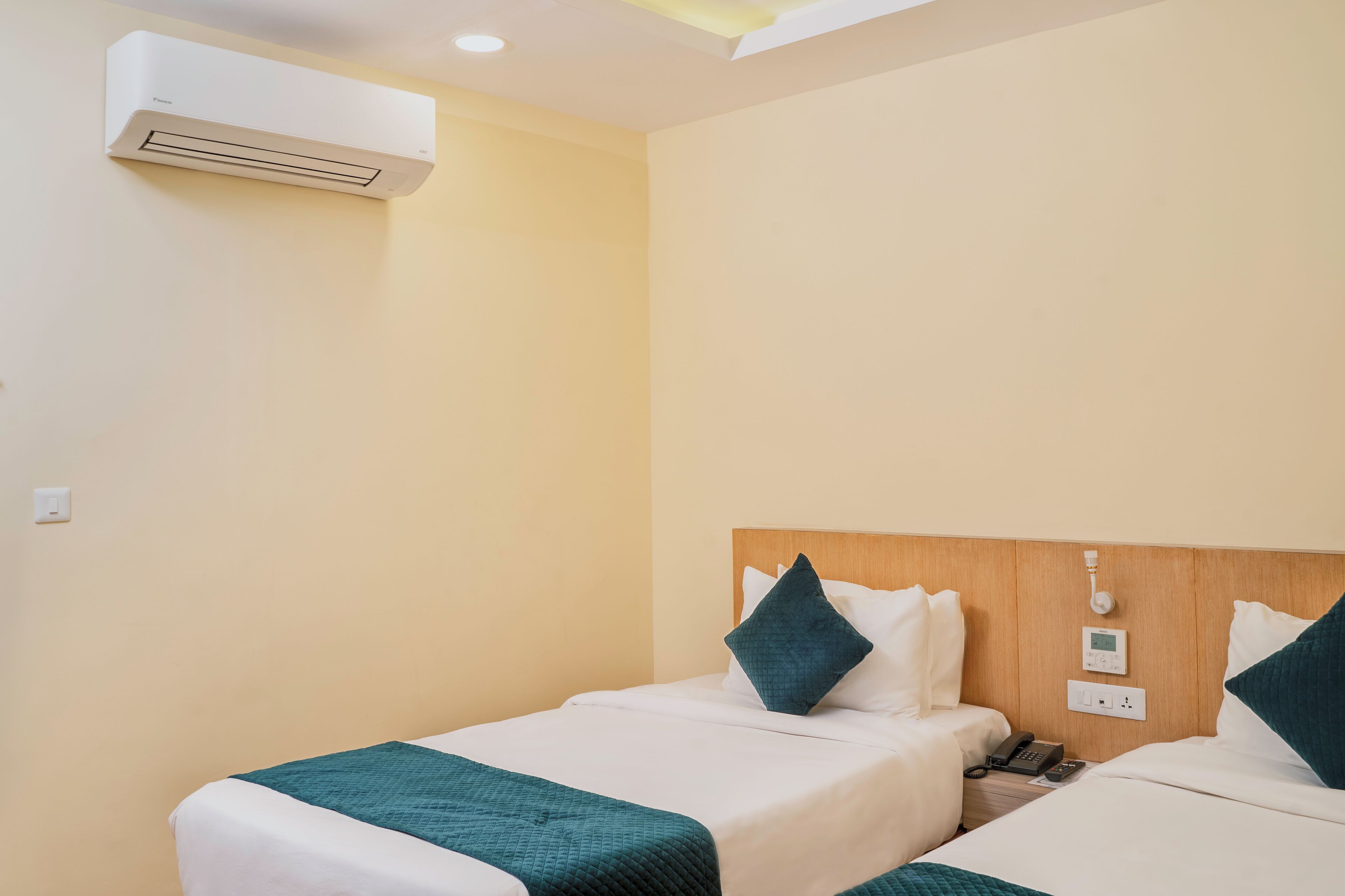 A hotel room with two beds, a wall-mounted air conditioner, a bedside telephone, and a power outlet.