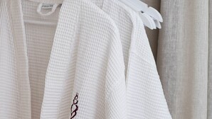 Una bata blanca colgada en un perchero blanco, con una etiqueta que dice "Coco-Ma", en una habitación con cortina de color neutro.