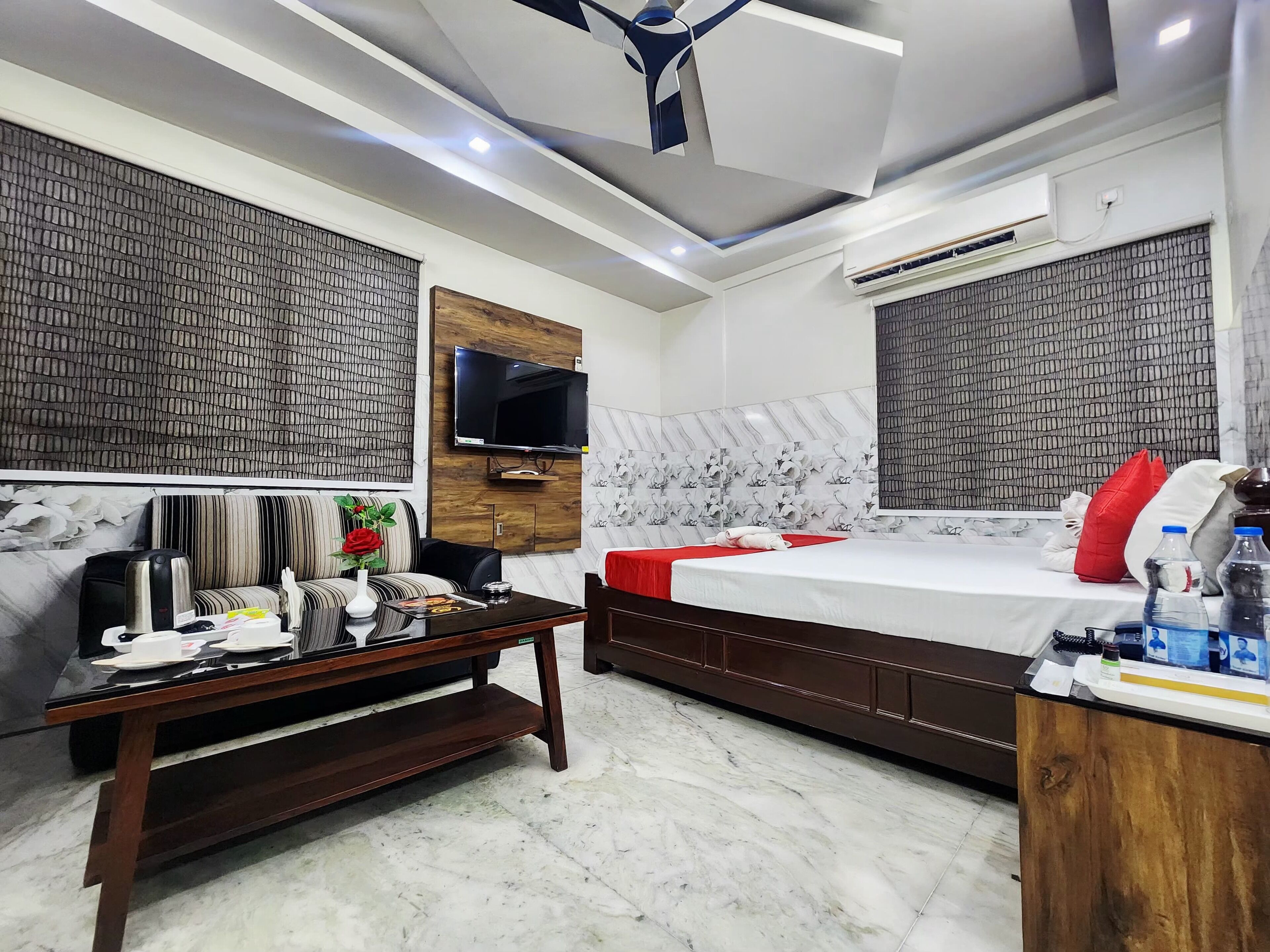 A hotel room with a bed, a sofa, a TV mounted on the wall, and an air conditioning unit.