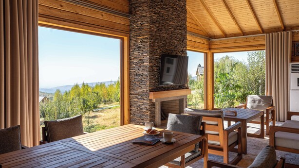 A cozy dining area with a stone fireplace, wooden furniture, and large windows offering a scenic view. - Silver Valley (Kumushkan)