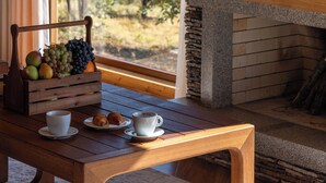 A wooden table with a fruit basket, cups, and pastries, a fireplace, and a view of trees through a window. - Silver Valley (Kumushkan)