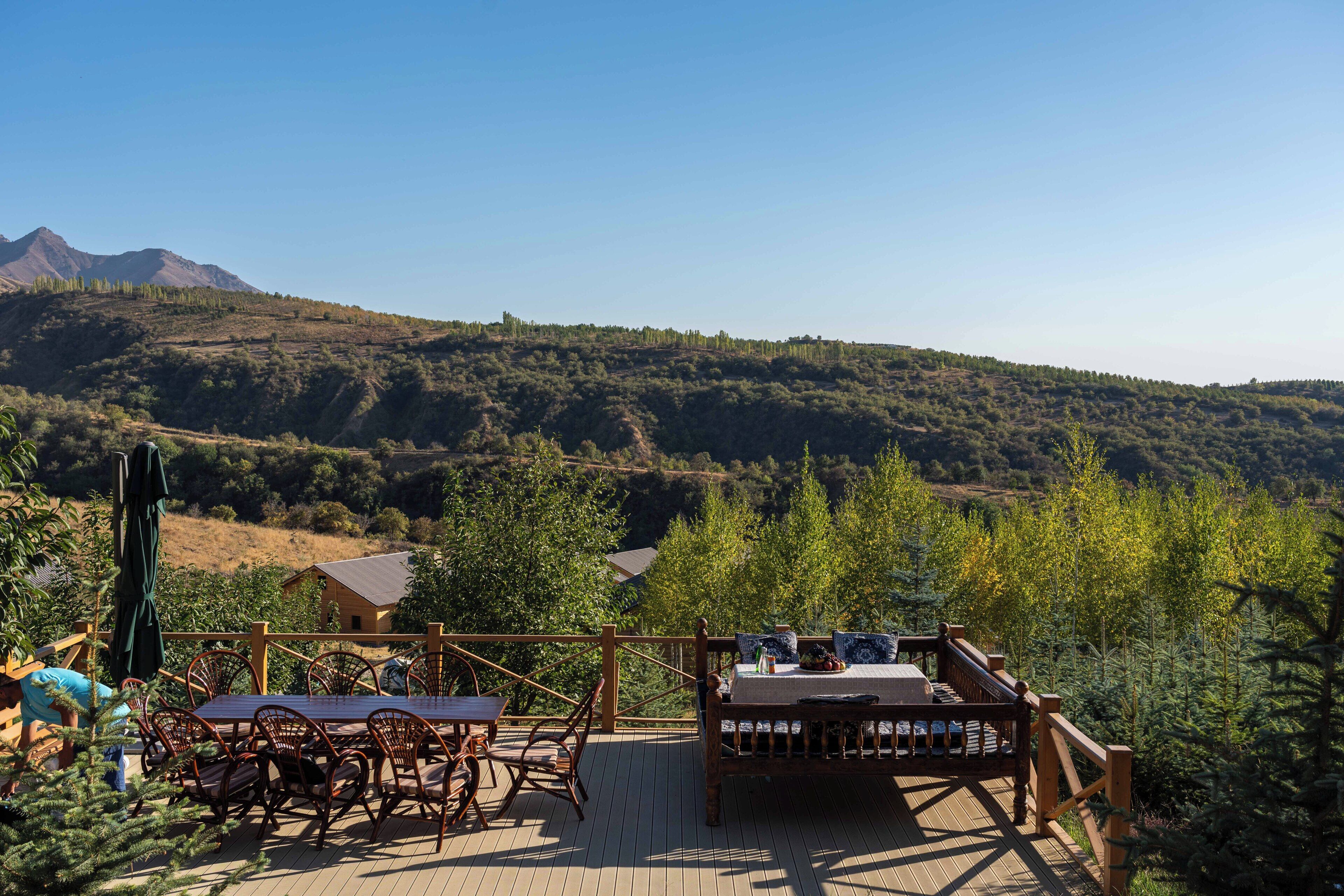 A wooden deck with a table and chairs, overlooking a mountainous landscape with trees and a house in the distance.