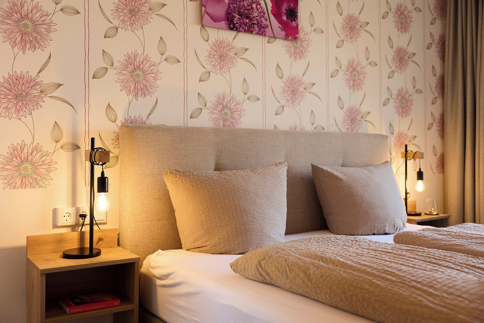 A bedroom with a bed, pillows, a nightstand, a lamp, and a wall with floral wallpaper.