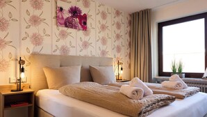A hotel room with a large bed, two bedside tables, a nightstand, a window with curtains, and a floral wallpaper design.