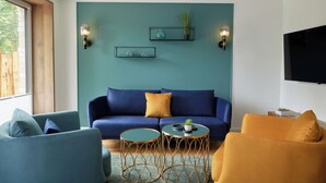 Una sala de estar moderna con una pared de acento color verde azulado, un sofá azul, un sillón amarillo y dos mesas redondas con patas de alambre dorado.
