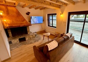 A cozy living room with a stone fireplace, a wooden ceiling, a large window, and a television.