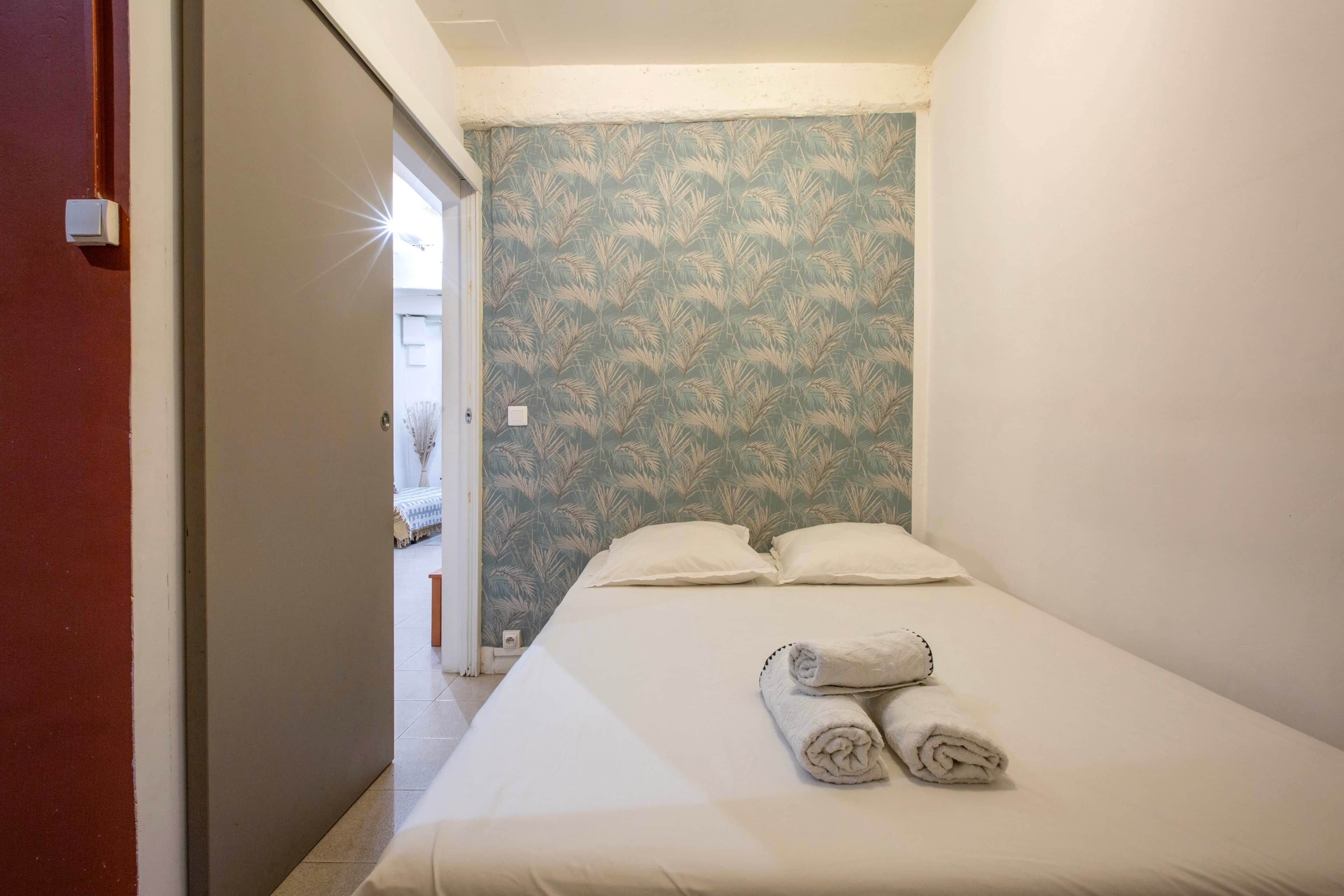 Een klein, net opgemaakt bed met twee kussens en twee opgerolde handdoeken, behang met een patroon, en een deur die leidt naar een andere kamer.