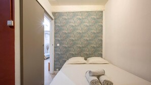 Una cama pequeña y ordenada con dos almohadas y dos toallas enrolladas, un papel tapiz estampado, y una puerta visible que da a otra habitación.