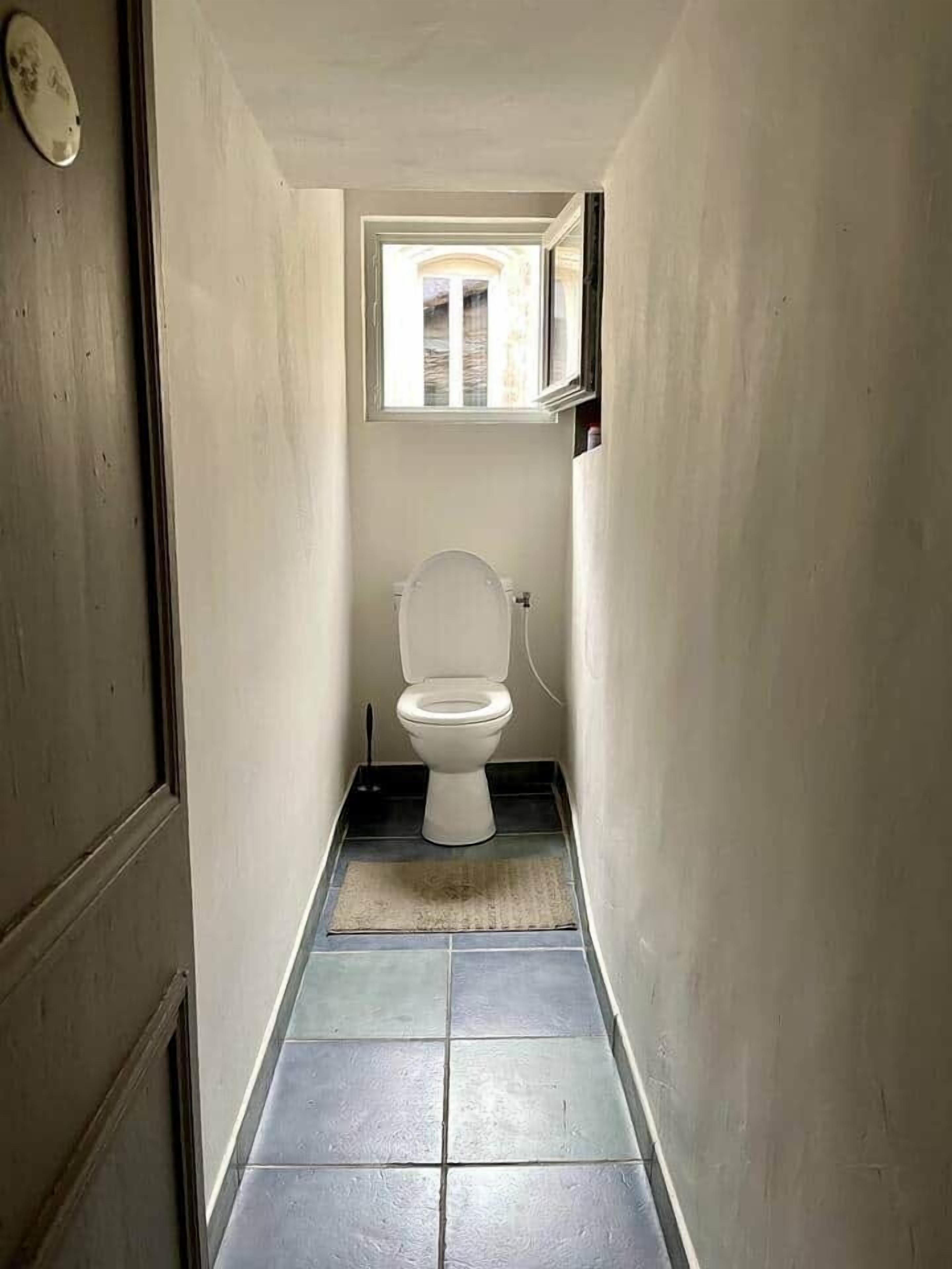 A narrow hallway with a toilet at the end, a small window above it, and a tiled floor.