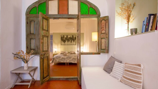 A room with a bed, a small table with a vase, and a mirror reflecting a bed and a wall with a decorative piece.