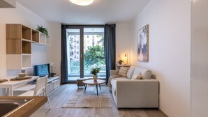 Un appartement moderne comprenant une cuisine, un coin repas et un salon. Il dispose d’une grande fenêtre offrant une vue sur les immeubles et les arbres.