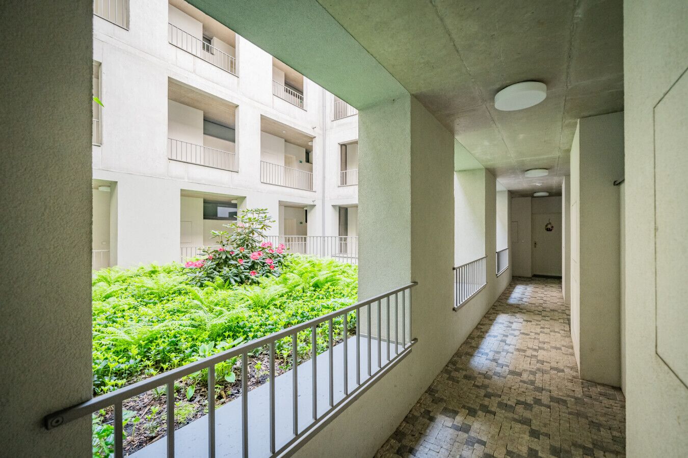 A corridor with a balcony overlooking a garden with green plants and pink flowers.
