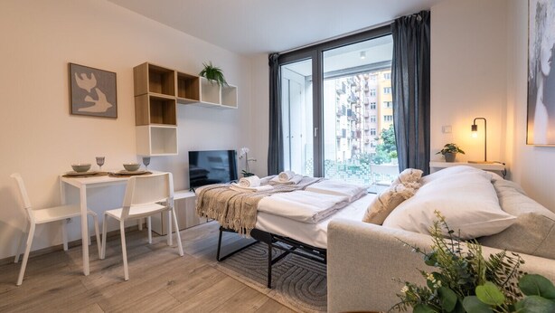 Une chambre moderne et bien éclairée, équipée d’un lit, d’une petite table avec des chaises, d’une télévision et d’une grande fenêtre avec des rideaux.