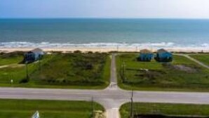 An aerial view of a coastal area with a road leading to a beach. There are several structures with blue roofs, possibly beach huts or small buildings, on grassy plots. - Cozy Seaside Cottage Escape (Port Bolivar)