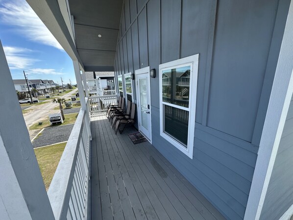 A covered porch with white railings, a series of windows, and a view of the street and other buildings in the background. - Cozy Seaside Cottage Escape (Port Bolivar)