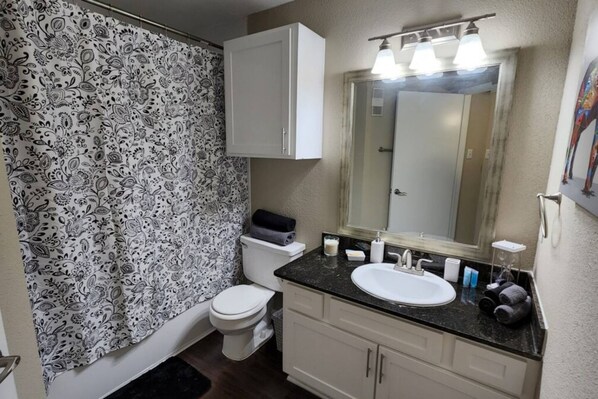 A bathroom with a patterned shower curtain, a toilet, a sink with a mirror, and a cabinet. - Medical Center 2BR With Resort Amenities (Houston)