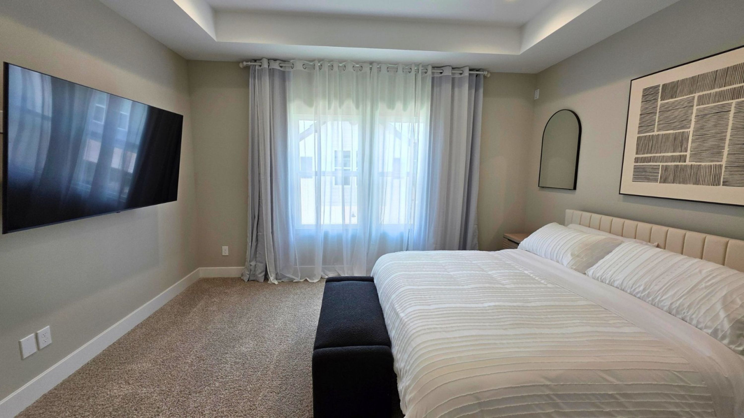 A bedroom with a large bed, a flat-screen TV mounted on the wall, a window with sheer curtains, and a framed abstract artwork above the bed.