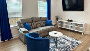 Un salon moderne avec un canapé, un fauteuil bleu, une table basse, un téléviseur et une étagère blanche.