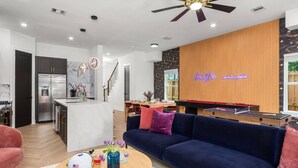 A modern living room with a dark blue sofa, a coffee table with a helmet and flowers, a ping pong table, and a wooden accent wall with a neon sign. - Family Retreat Near NRG Med Center (Houston)