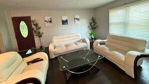 Ein Wohnzimmer mit zwei weißen Ledersofas, einem Glastisch und einem dunklen Holzboden. An den Wänden hängen Bilder in Rahmen und in einer Glasvase steht eine Pflanze.