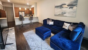 A modern living room with a blue sectional sofa, a white rug, and a kitchen in the background. - Elite Downtown Poolside Oasis (Houston)