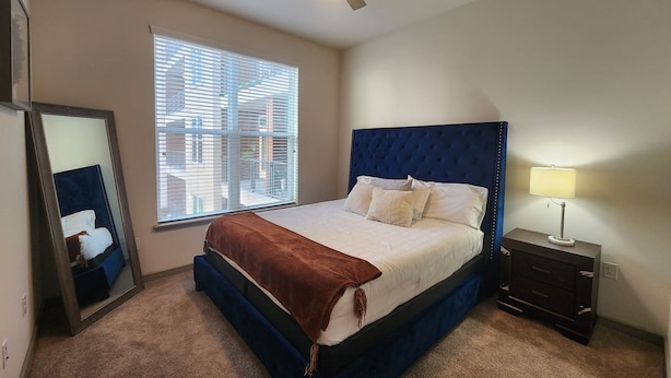 A bedroom with a bed, a ceiling fan, a nightstand with a lamp, and a window with blinds. - Elite Downtown Poolside Oasis (Houston)