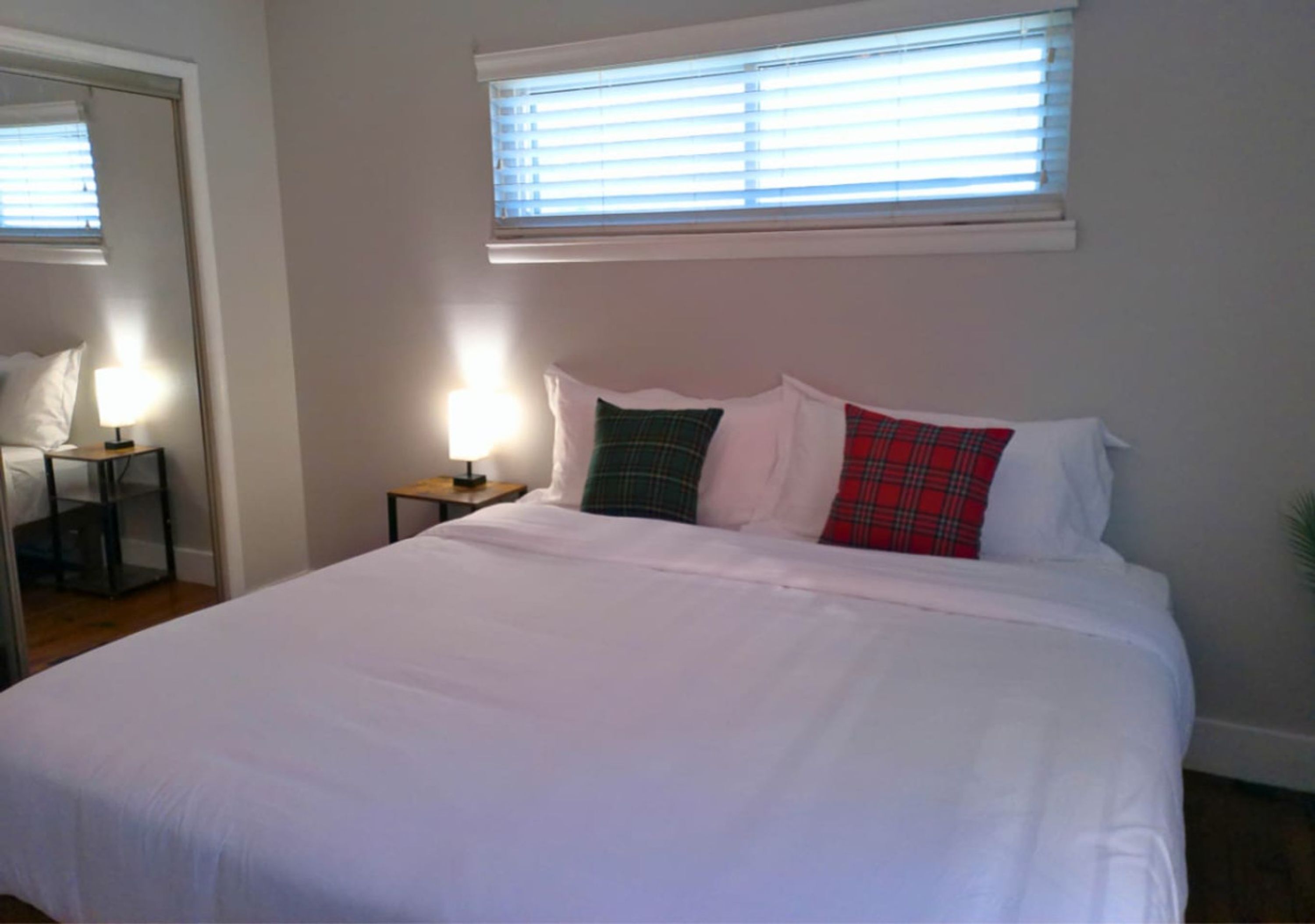 Een net opgemaakt bed met wit beddengoed en geruite kussens, een nachtkastje met een lamp, en een raam met jaloezieën.