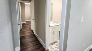 Un couloir doté d’un plancher en bois mène à une salle de bain avec un lavabo et des meubles de rangement blancs.
