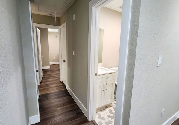 A hallway with a wooden floor leading to a bathroom with a white sink and cabinets.