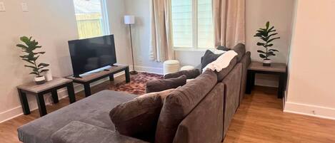 A living room with a grey sectional sofa, a flat-screen TV on a stand, a small table with a plant, and a window with curtains.