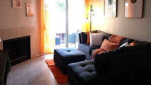Un salon comprenant un canapé, un pouf, une cheminée et une vue sur l’extérieur, visible à travers une porte vitrée.