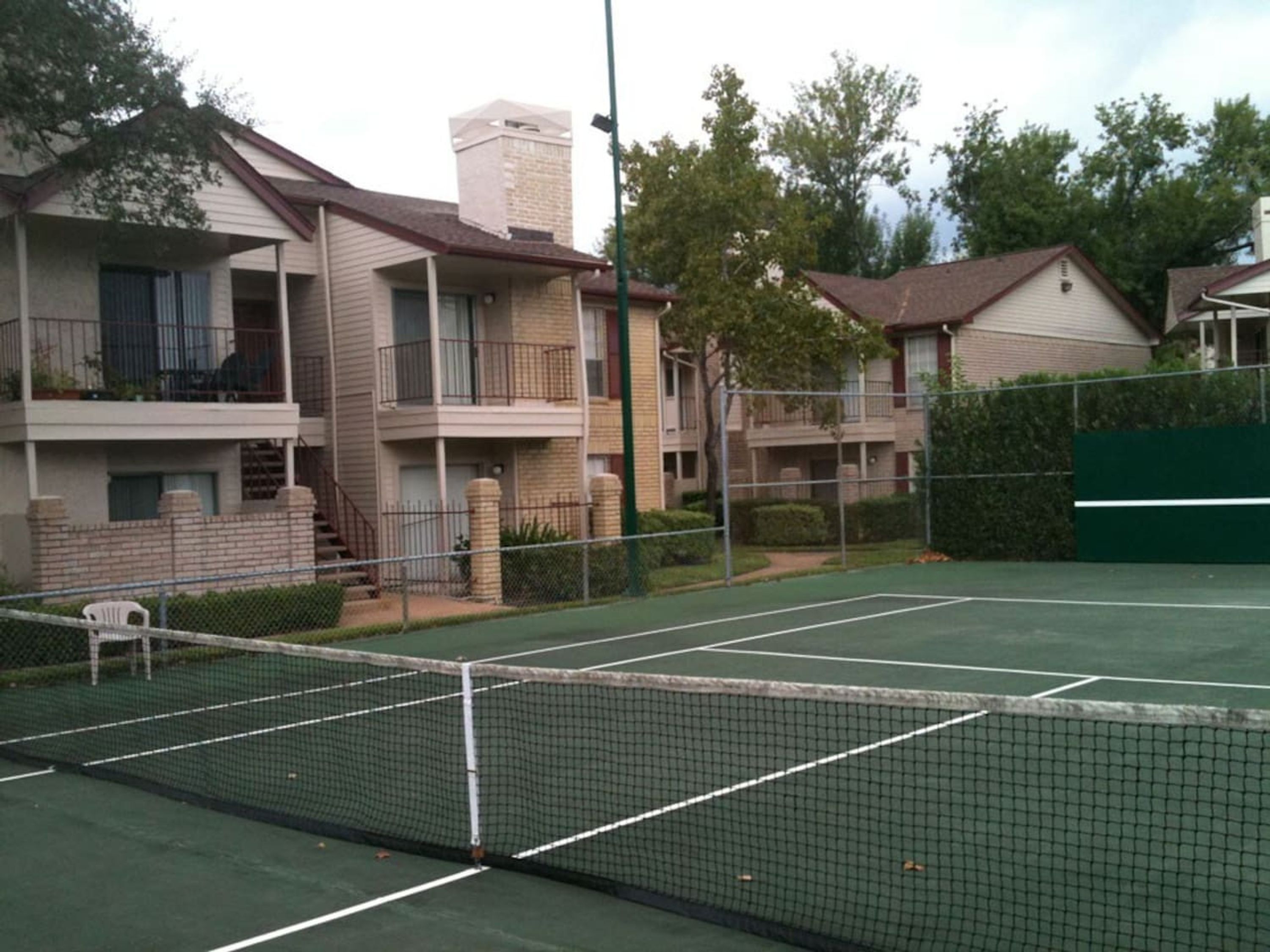 En tennisbana med ett nät i mitten, omgiven av bostadshus med balkonger och ett grönt staket.