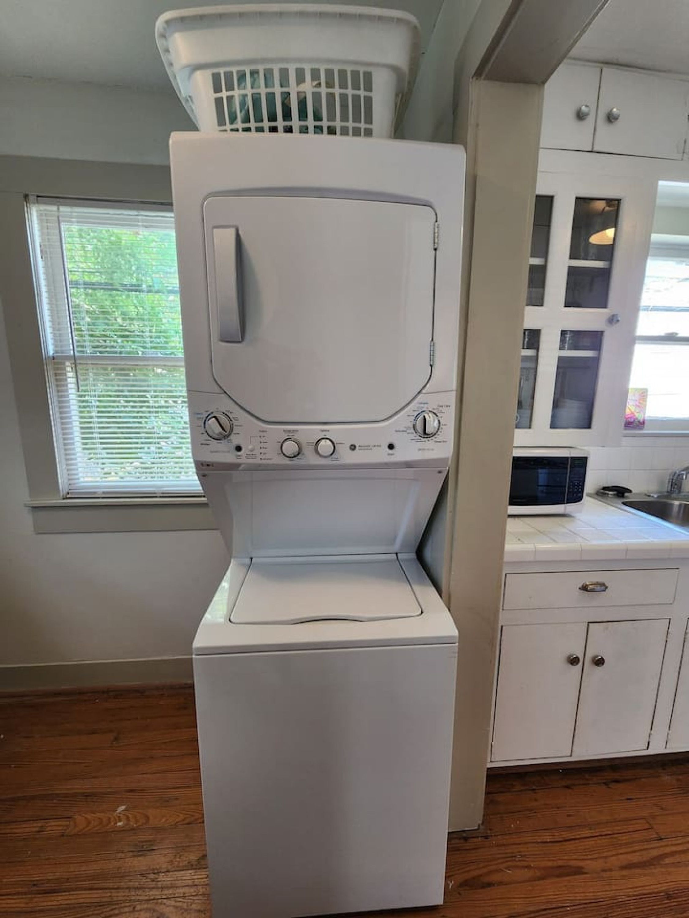 A compact washer and dryer unit in a kitchen with white cabinets and a window with blinds.