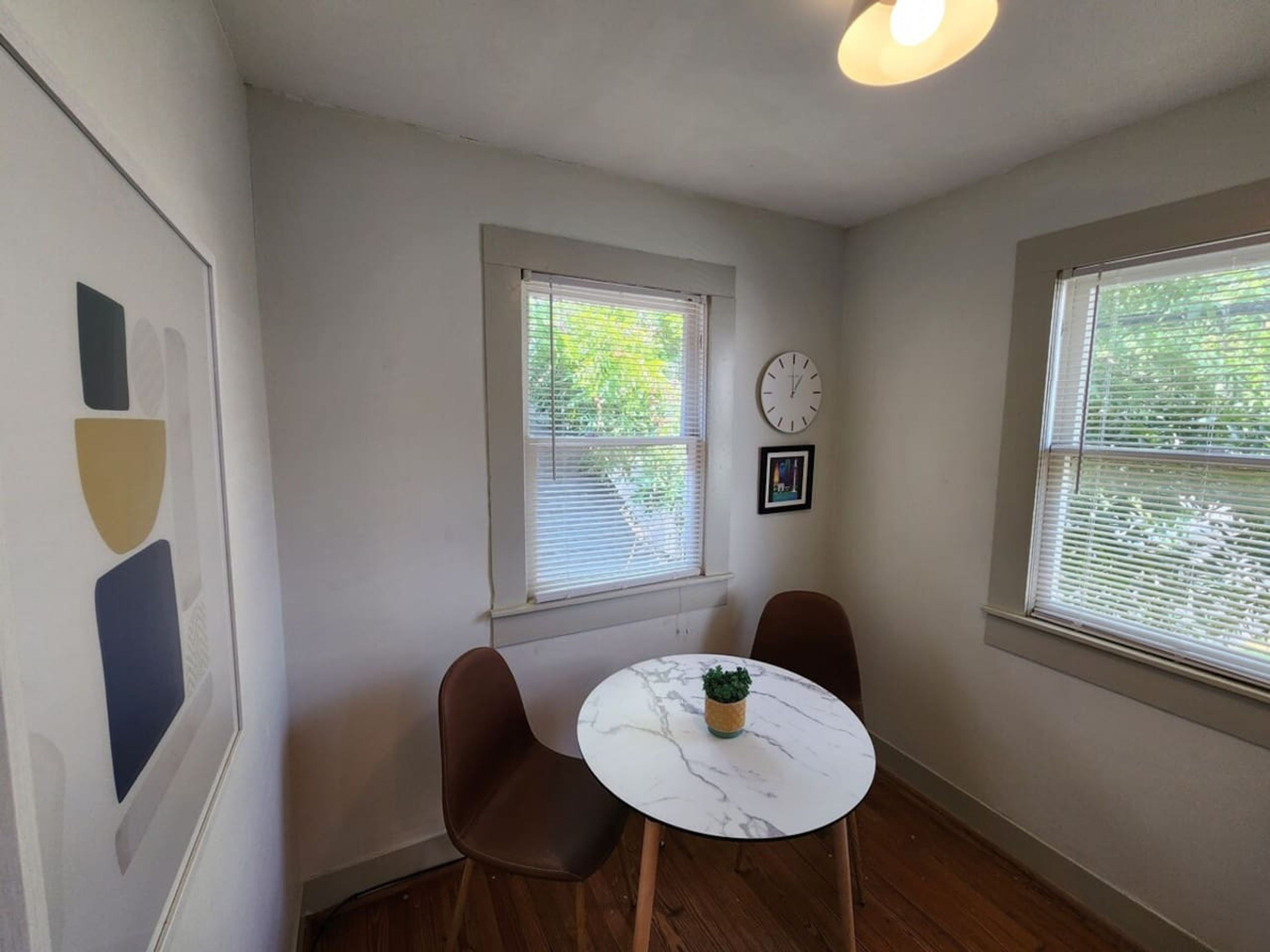 A small room with two windows, a clock, and a framed picture on the wall.