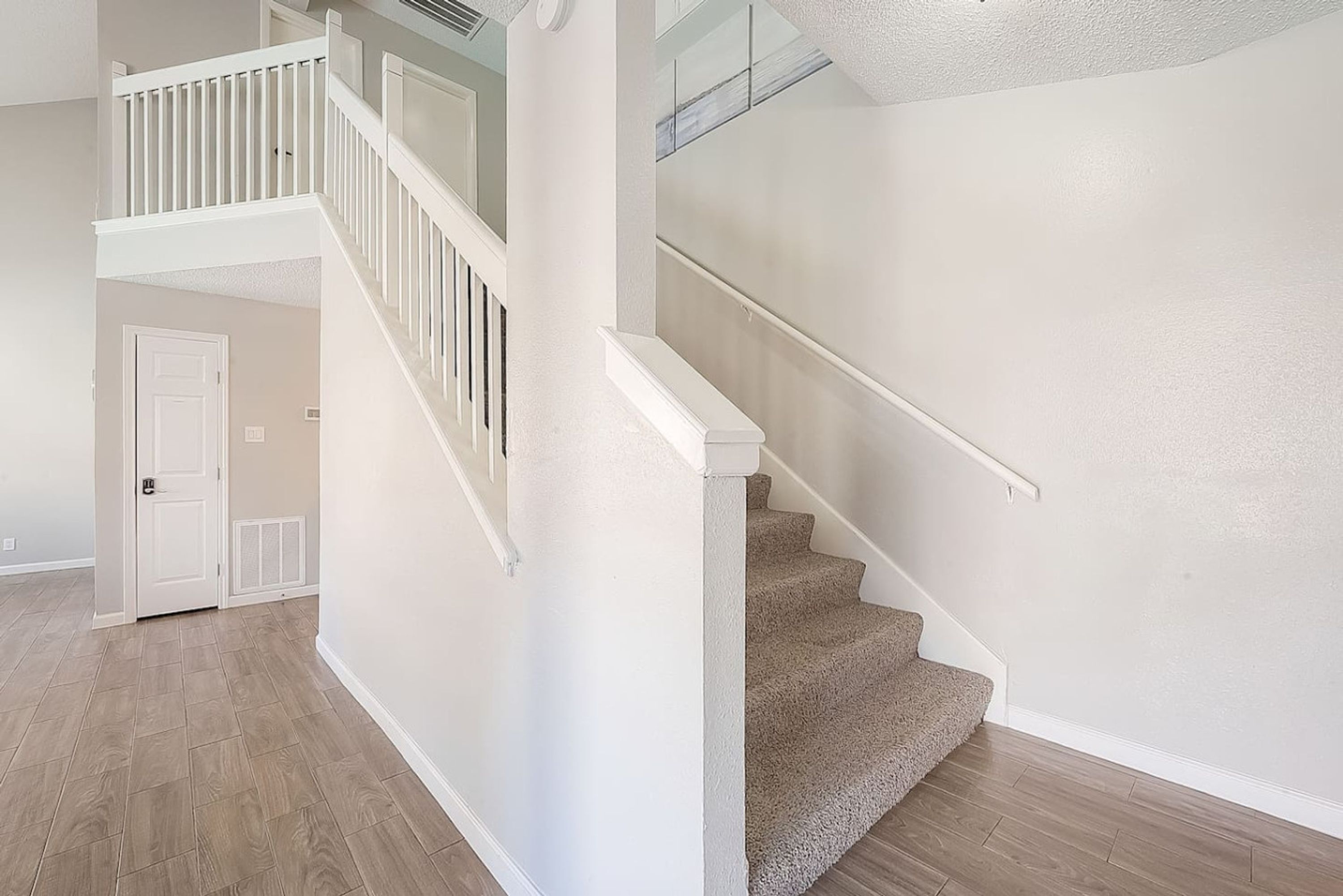 Un escalier avec une rampe blanche mène à un palier. Les murs sont peints dans une couleur claire et le sol est recouvert de parquet de couleur marron clair.