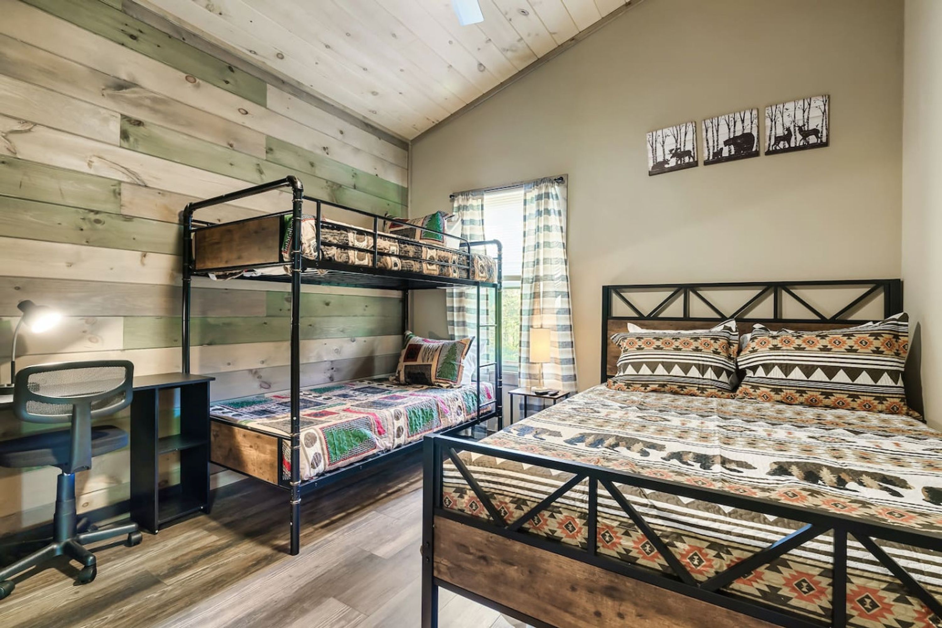 A room with a bunk bed, a desk, and a chair. There is a window with curtains and a wall-mounted artwork.