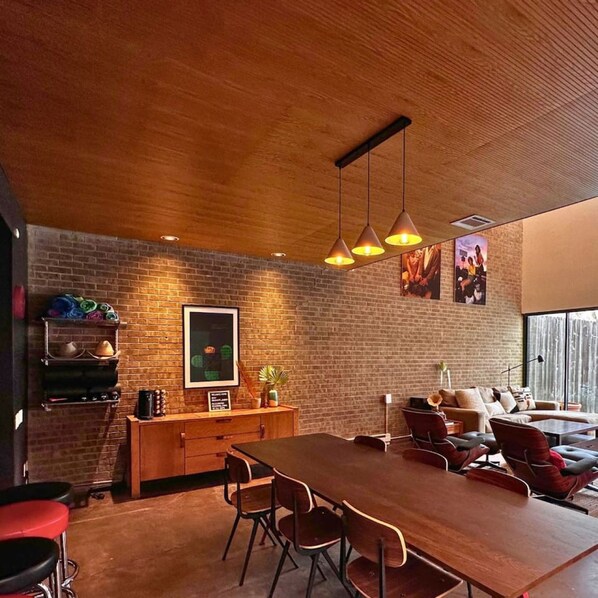 A modern dining area with a brick wall, wooden ceiling, and pendant lighting. There is a wooden table with chairs, a bar area with stools, and a sofa in the background.
