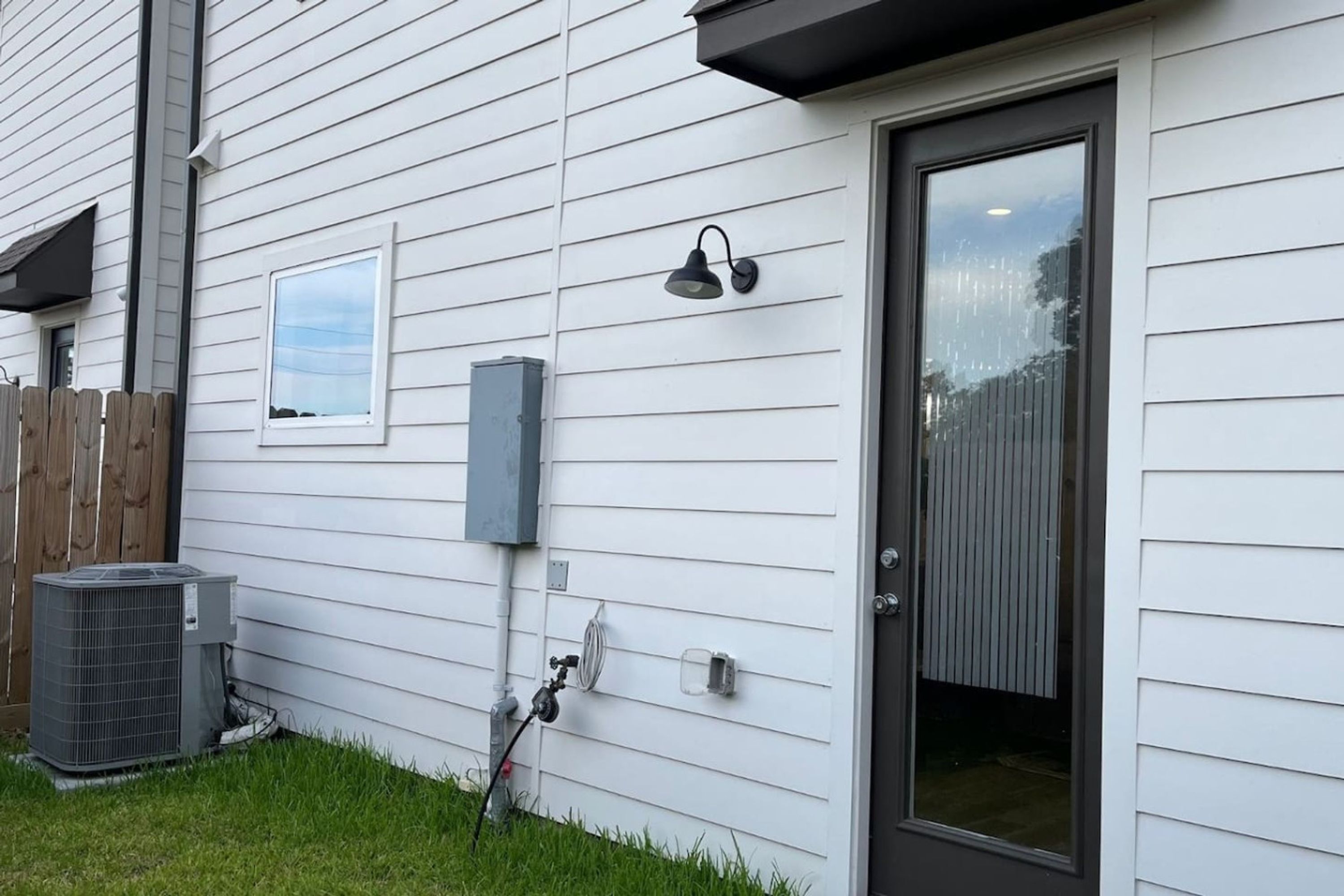 A house with white siding, a front door, a window, a wall-mounted light fixture, an air conditioning unit, and a wooden fence.