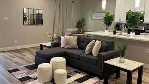 Uma sala de estar moderna com um sofá seccional, pufes, uma mesa de centro e uma área de cozinha com armários e eletrodomésticos brancos.