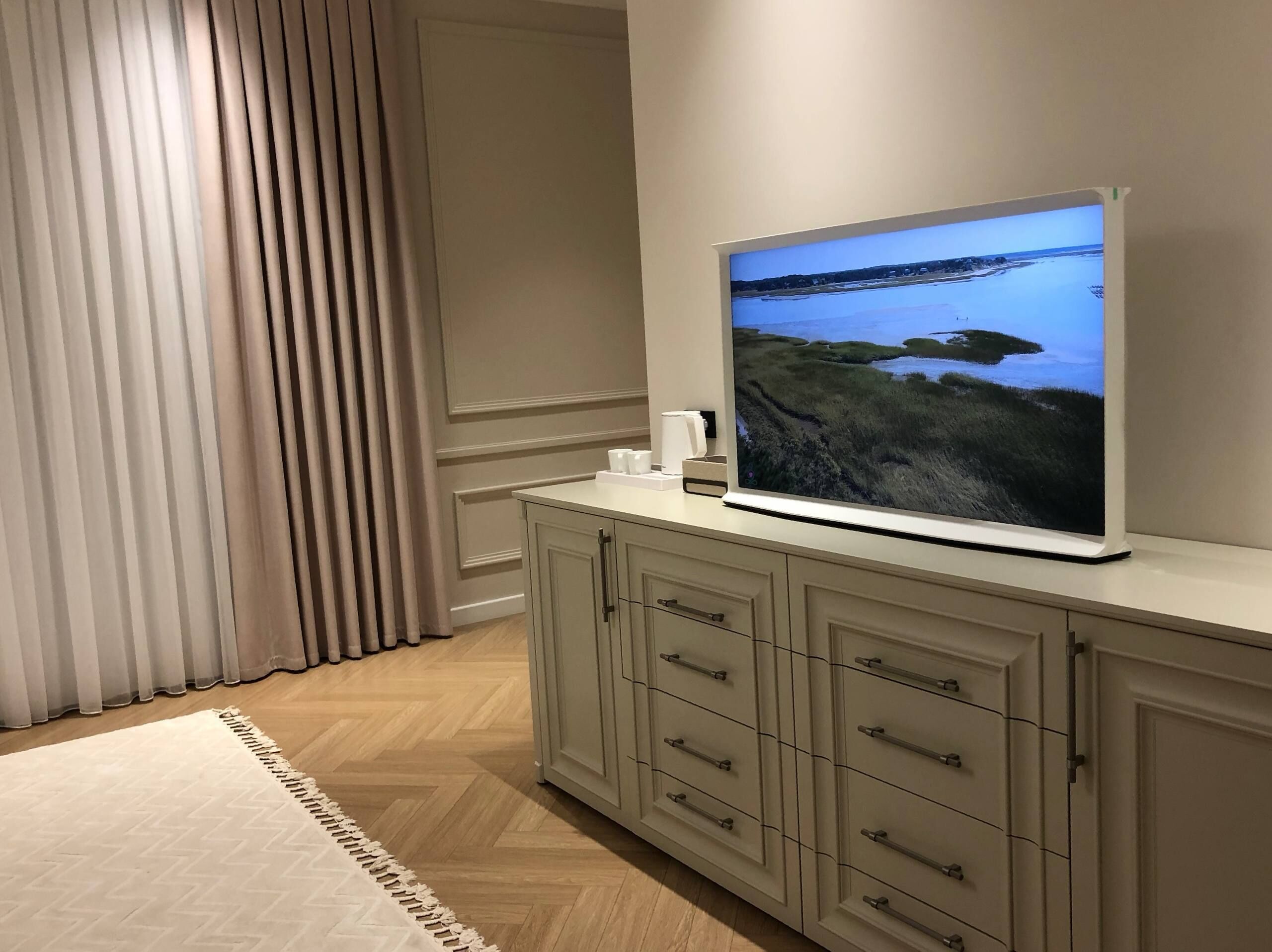 Ein modernes Wohnzimmer mit einem an einer Kommode befestigten Flachbildfernseher, einem großen Fenster mit Vorhängen und einem Teppich mit Zickzackmuster.
